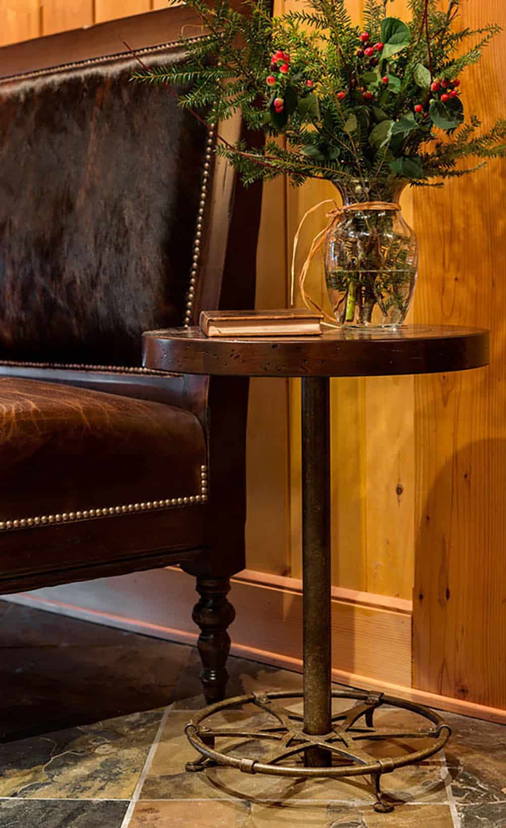 Round distressed wood side table on an iron base beside a cowhide and leather bench, with a vase of evergreens and red berries