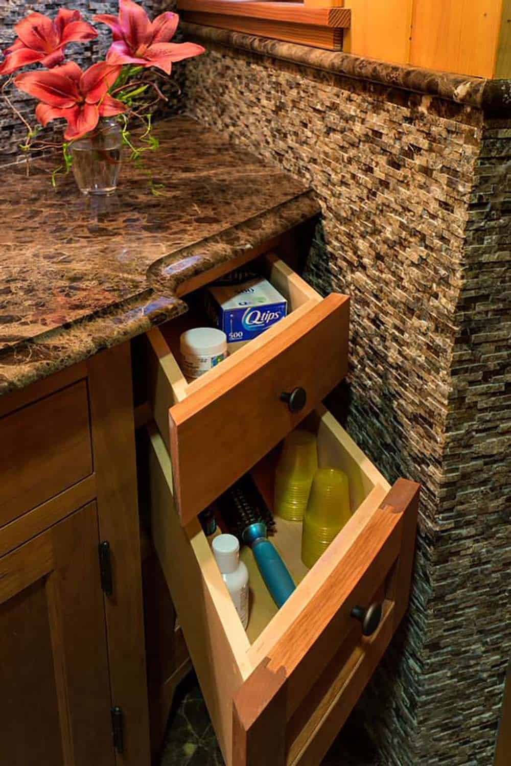 Open cherry vanity drawers beneath a brown marble countertop beside a stacked stone mosaic wall
