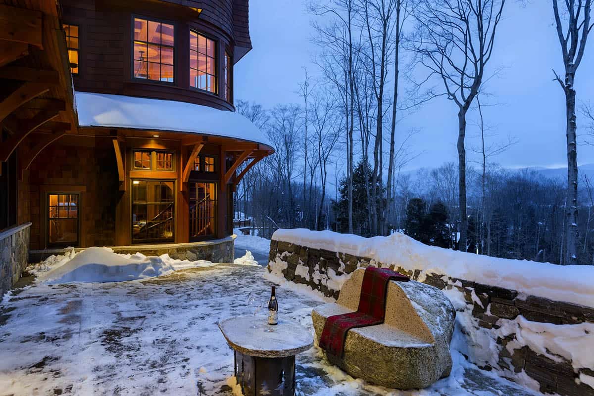 Snowy stone terrace beside the turret at dusk, with a boulder bench draped in a red plaid throw and a wine bottle on a stone side table