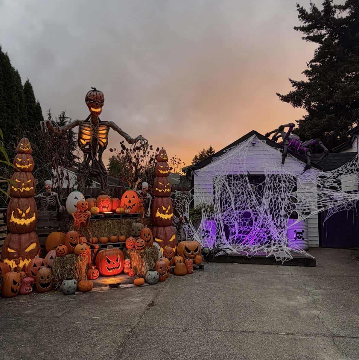 Glowing pumpkin towers and a giant pumpkin-headed skeleton at dusk beside a garage covered in webbing with a giant spider on the roof