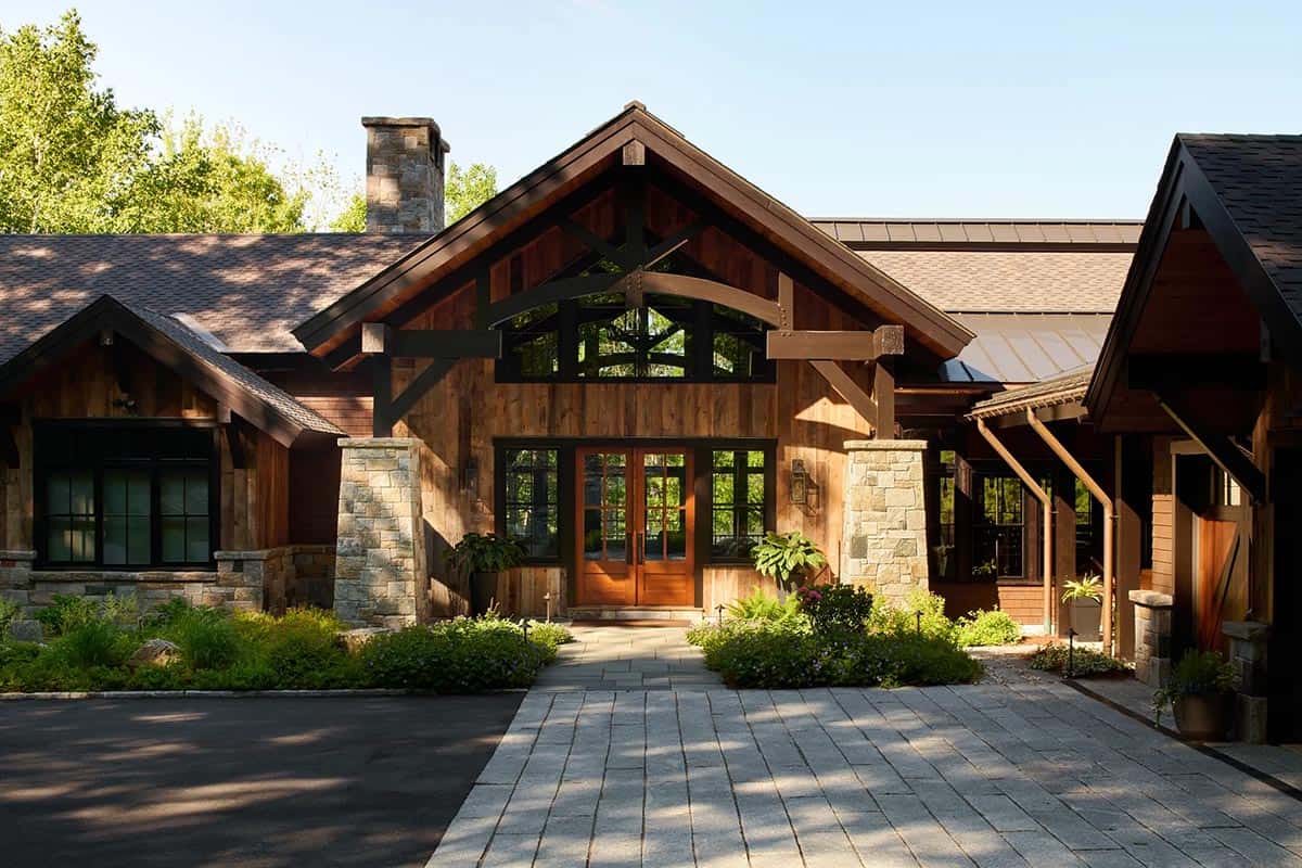 Rustic lodge-style home with a timber truss gable, weathered wood siding, stone piers and a granite paver drive in summer sun