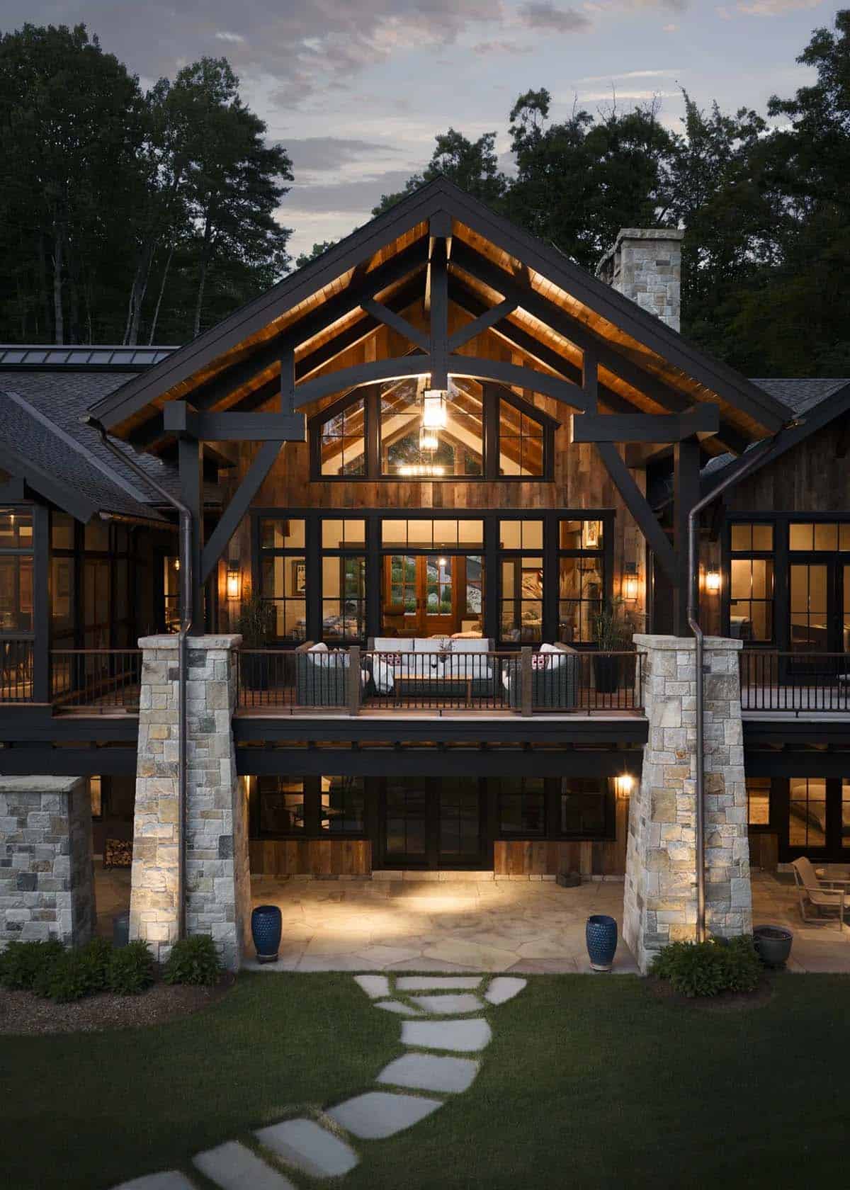 Timber truss gable glowing at twilight over an upper deck with wicker seating, stone piers and a flagstone patio with a stepping-stone path