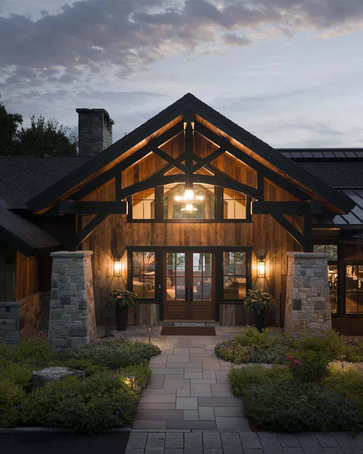 Front entry at dusk under a lit timber truss gable, with tapered stone piers, wood double doors and a stone walkway through garden beds