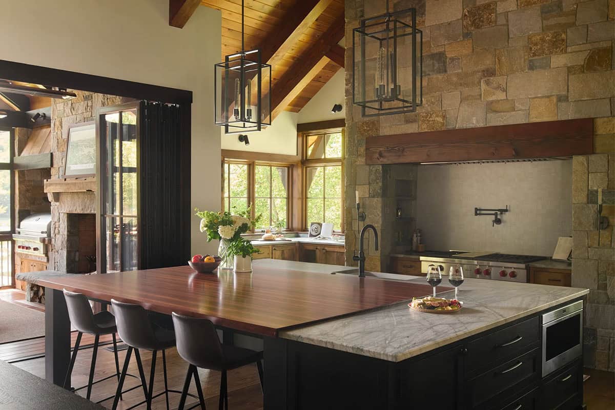 Kitchen with a dark island topped in wood and white marble, leather counter stools, lantern pendants and a range set into a stone wall