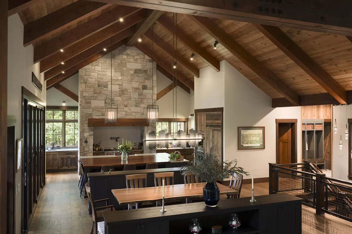 Vaulted great room looking past a live-edge dining table to the kitchen island, a stone range wall and a mudroom beyond