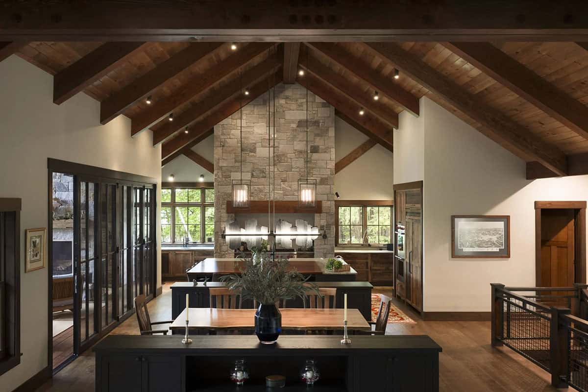 Great room with a vaulted wood ceiling and exposed beams, a dining table behind a dark console and a stone chimney wall over the kitchen
