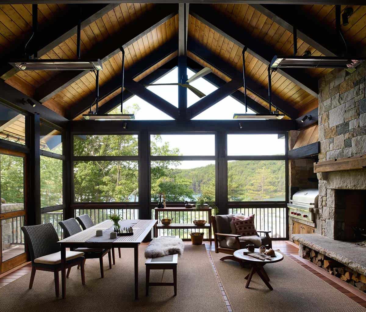 Screened porch with a vaulted wood ceiling and timber trusses, hanging heaters, a dining table, armchair and stone fireplace facing the lake