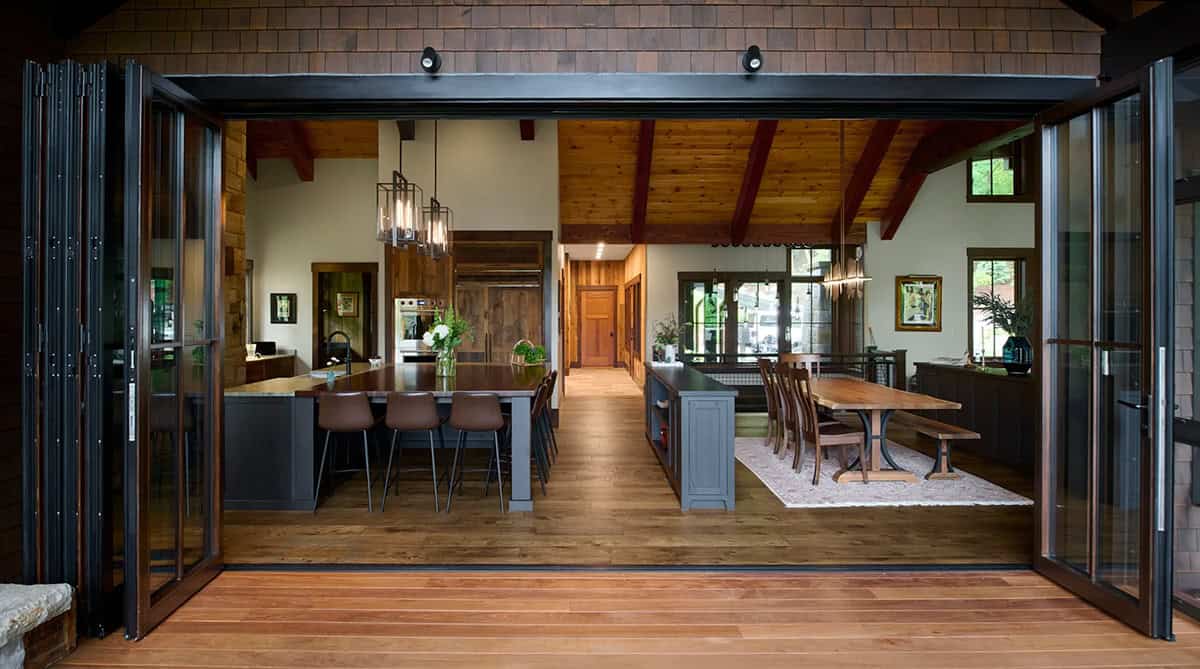 View from the porch through open folding glass doors into the kitchen and dining area with a long wood-topped island and trestle table