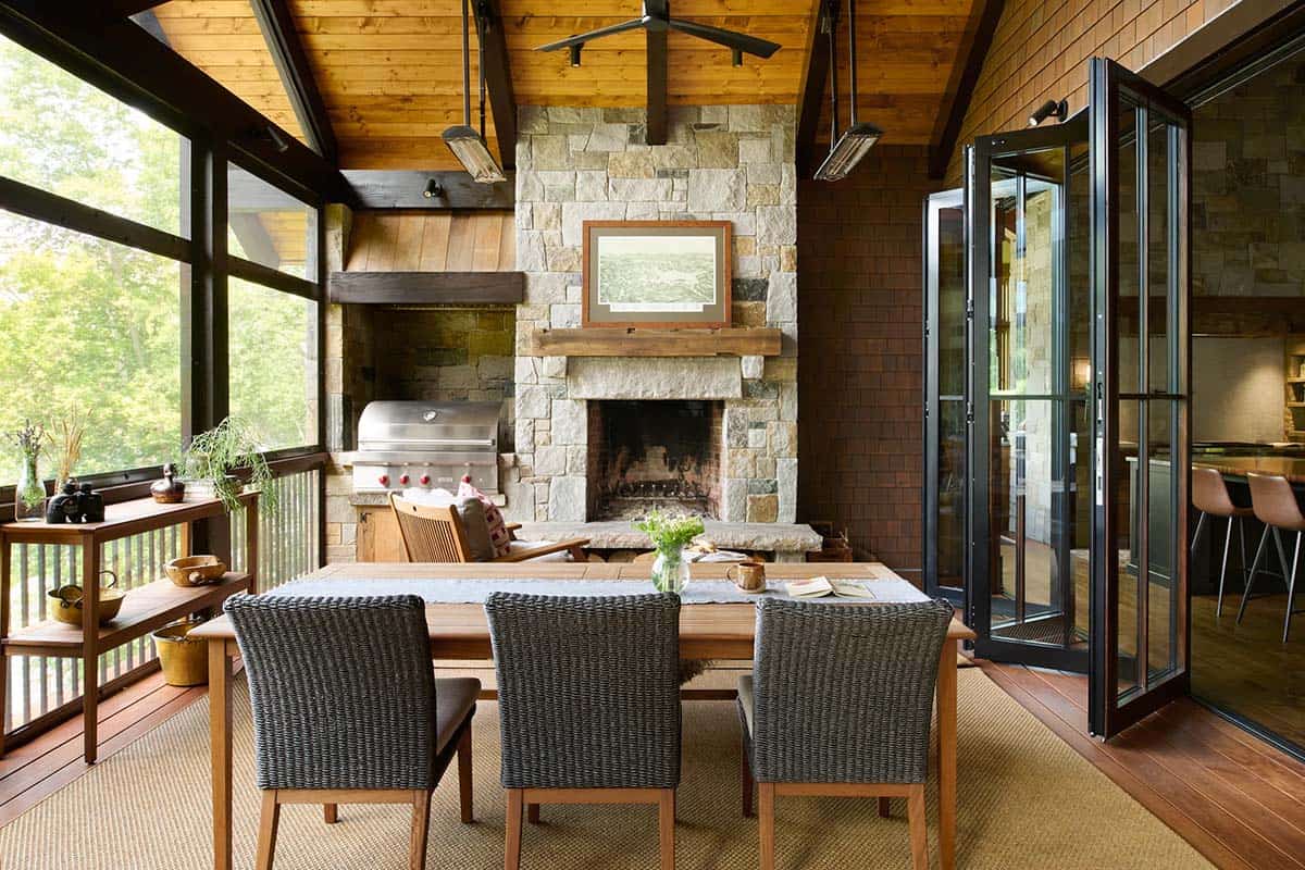Screened porch dining table with woven chairs before a stone fireplace and built-in grill, with folding doors open to the kitchen