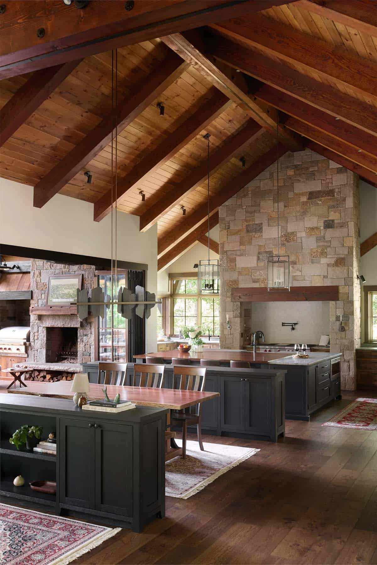 Great room with a vaulted wood ceiling, dark built-in cabinetry, a dining table and kitchen islands in front of a tall stone wall
