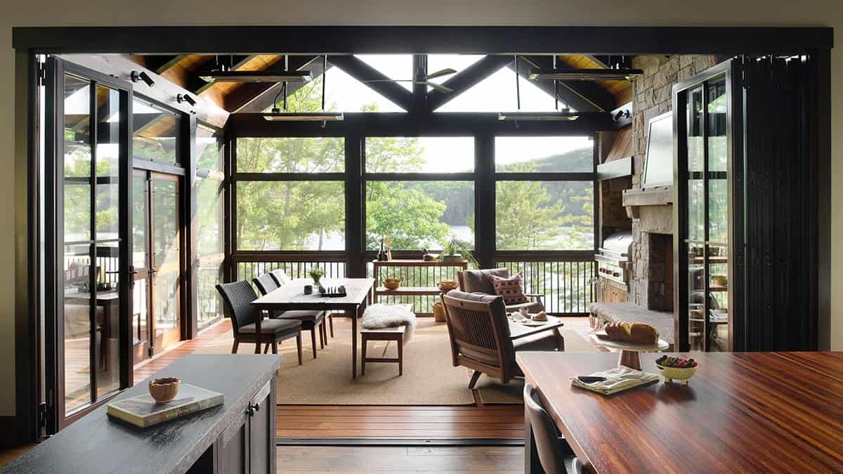Folding glass doors open from the kitchen to a screened porch with a dining table, armchairs, a stone fireplace and lake views