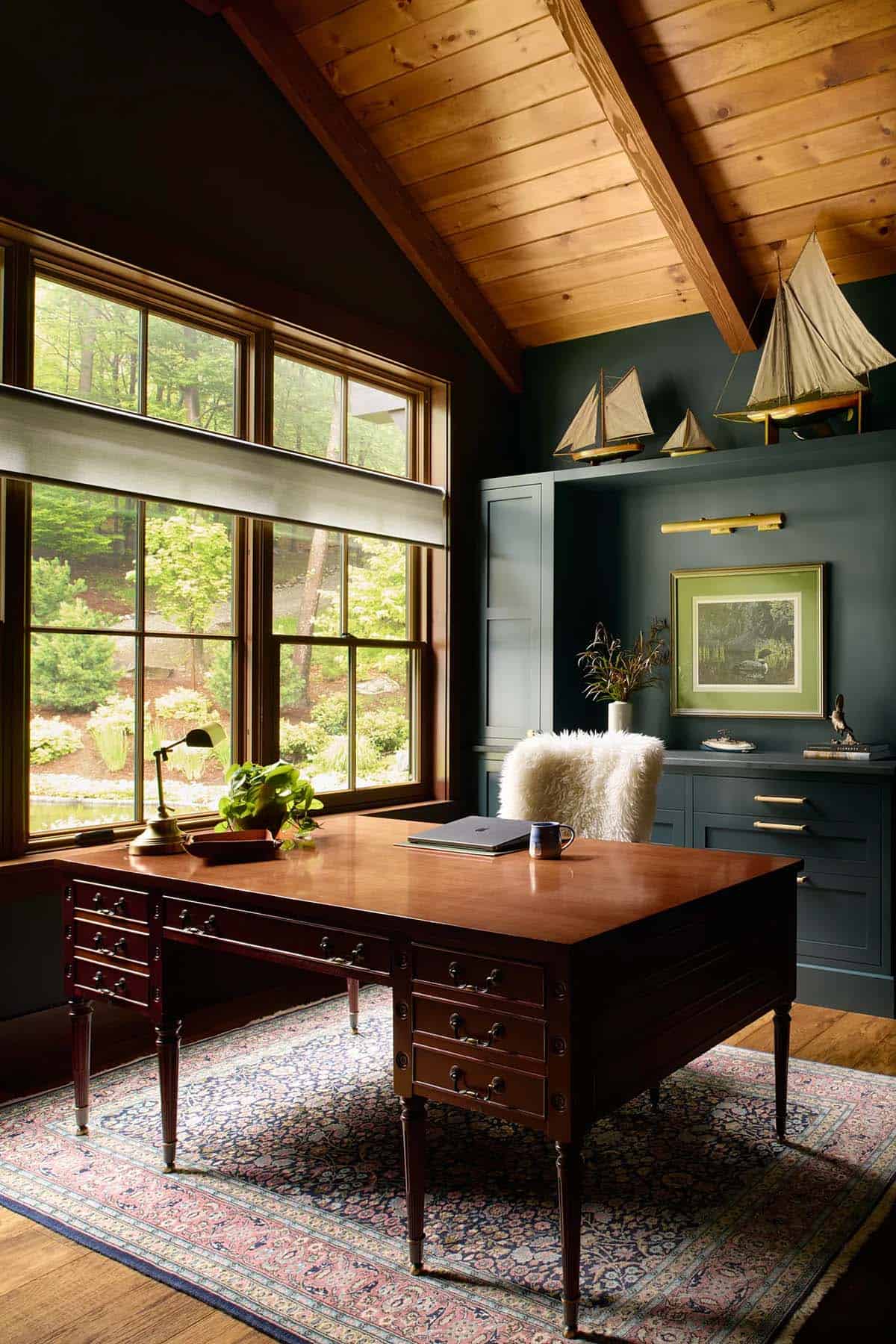 Home office with dark blue-green built-ins, model sailboats on a high shelf, an antique wood desk on a Persian rug and windows to the woods