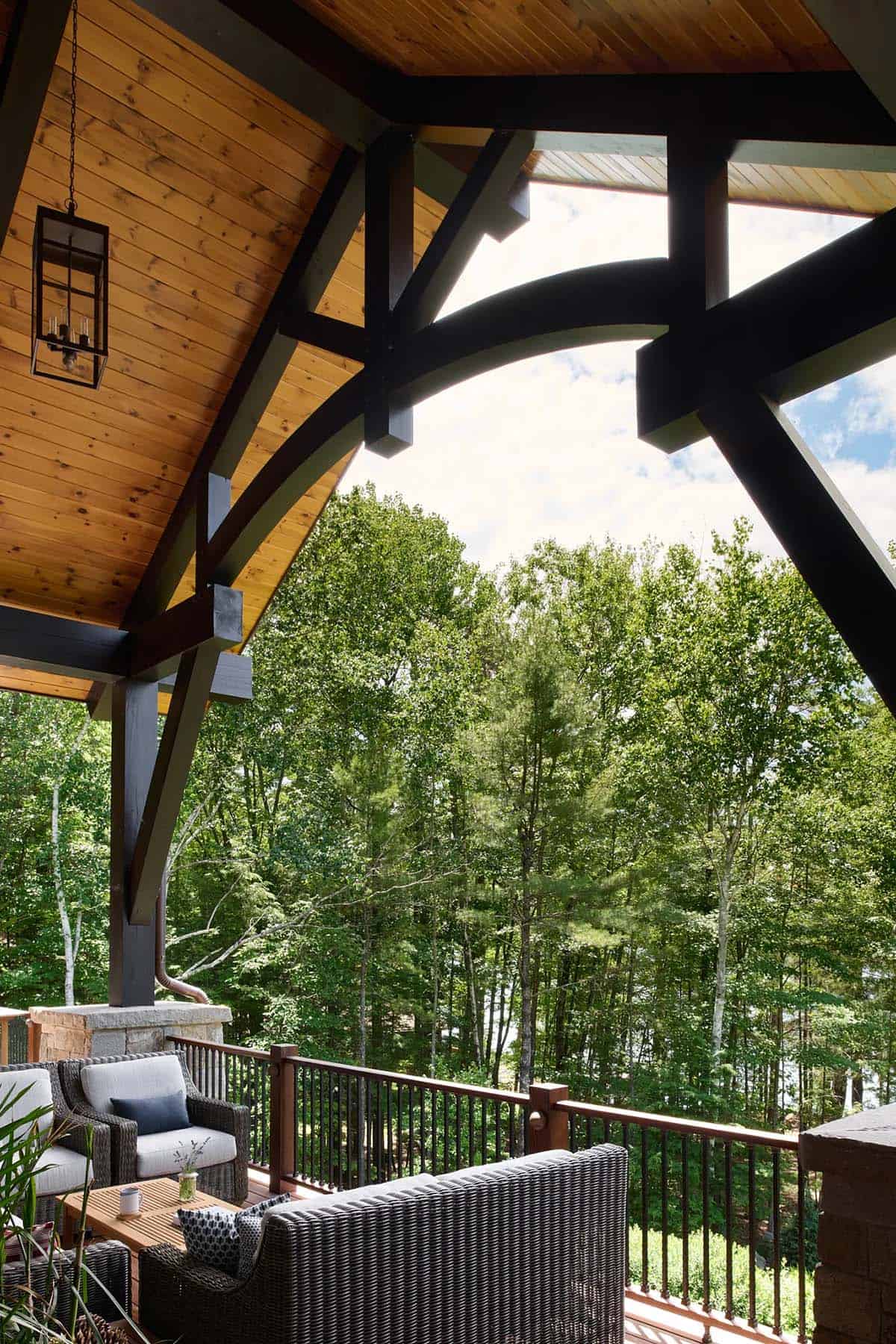 Covered deck with a wicker sofa and chairs beneath dark curved timber braces and a lantern, looking out to tall trees