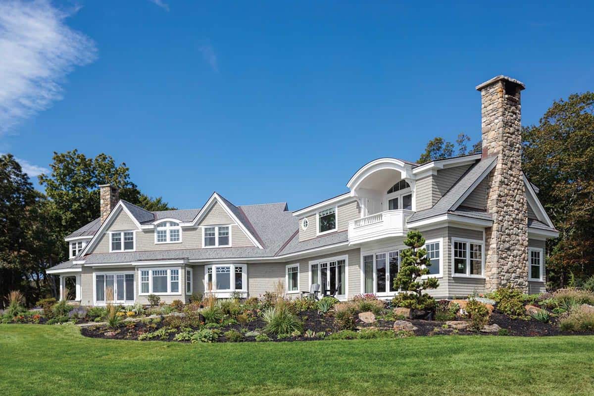 Side view of the shingle-style house with a tall stone chimney, bay windows, and a rock garden in front