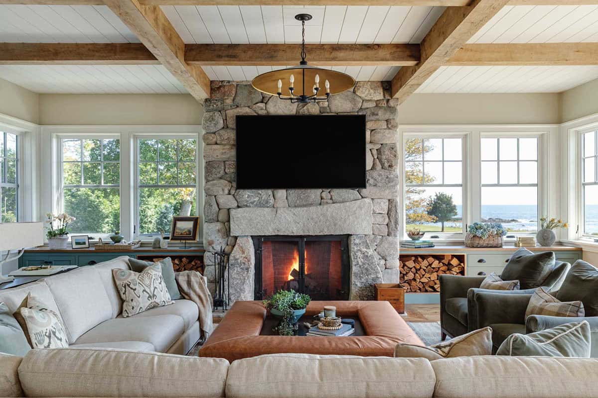 Living room with a fieldstone fireplace, beamed ceiling, sectional sofa, and leather ottoman