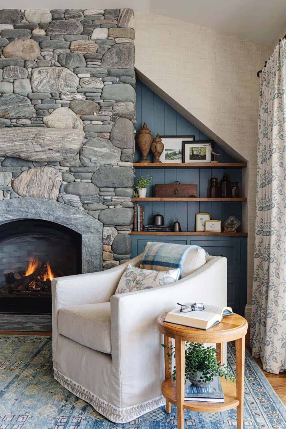 Reading corner with a gray stone fireplace, blue built-in shelves, a slipcovered armchair, and a patterned rug