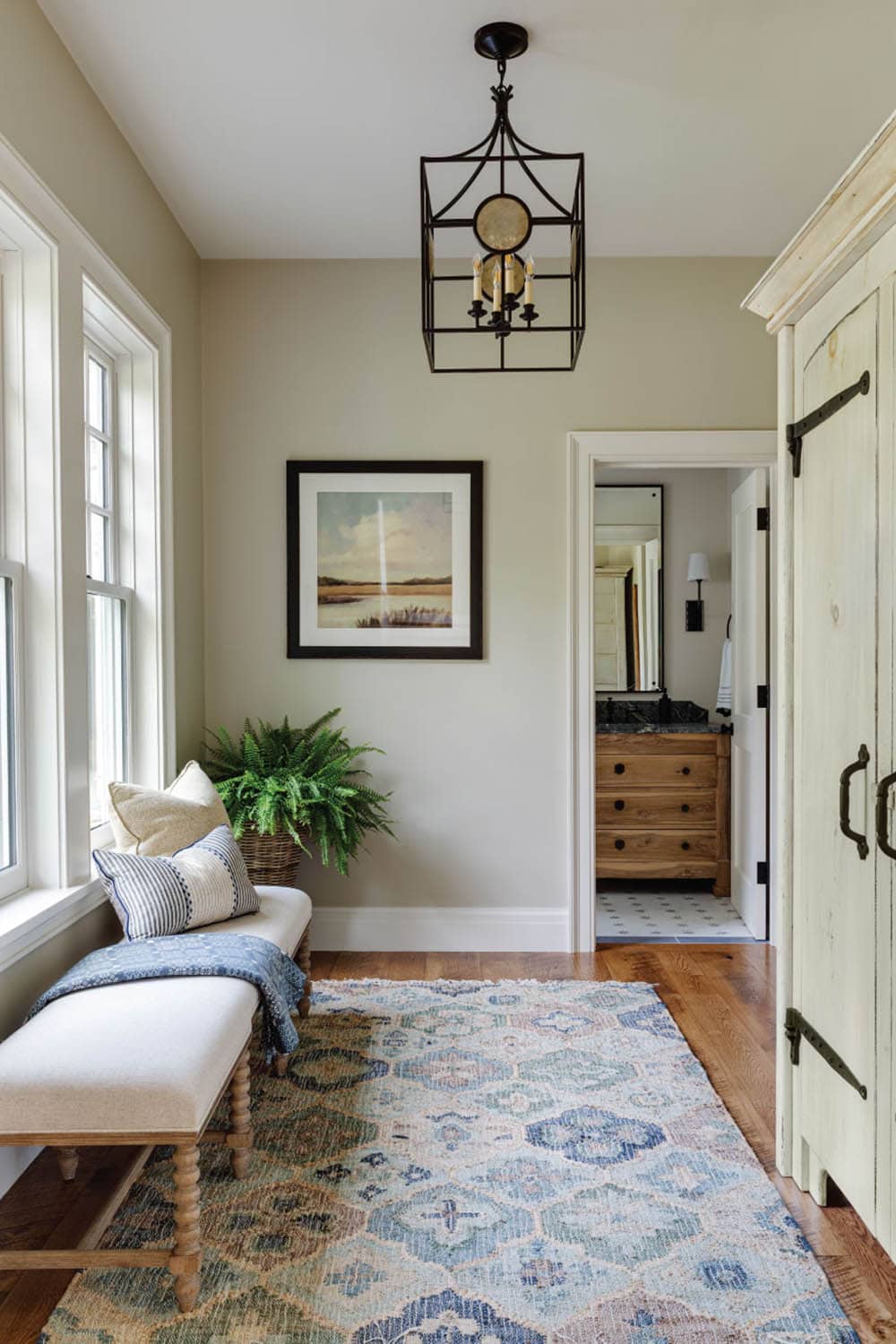 Dressing area with an upholstered bench, a whitewashed armoire, and a lantern pendant, with a vanity visible through the doorway