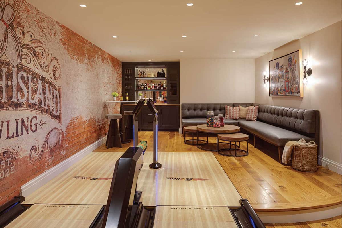 Bowling alley seen from the lane, with a dark bar cabinet, tufted leather banquette, and round coffee tables