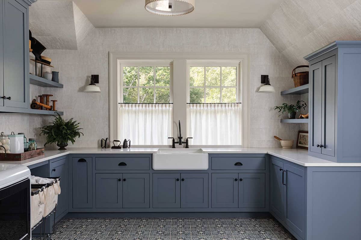 Laundry room with blue cabinets, a white farmhouse sink under a window with café curtains, and patterned floor tile