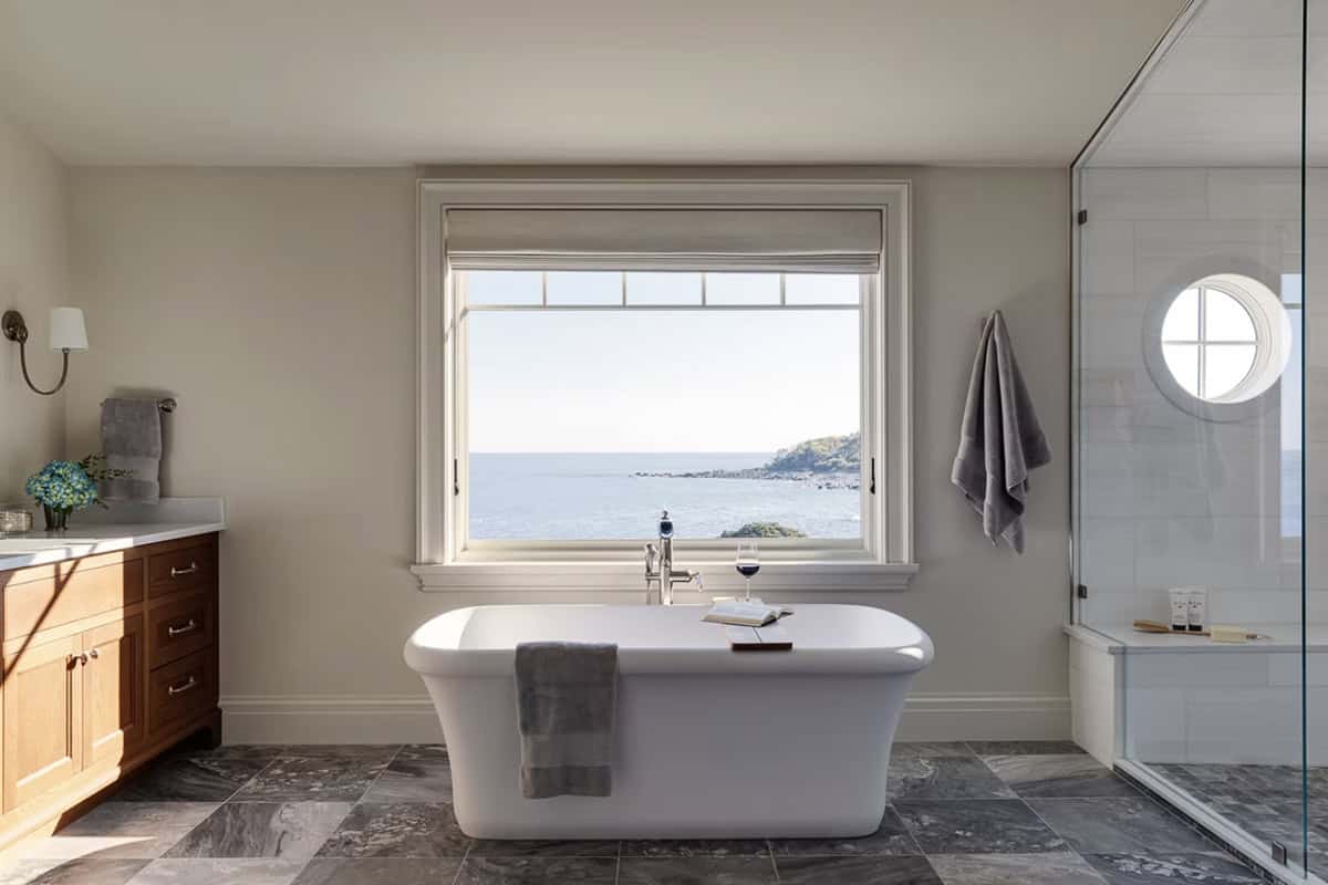 Bathroom with a freestanding tub beneath a large window overlooking the ocean, and a glass shower with a round window