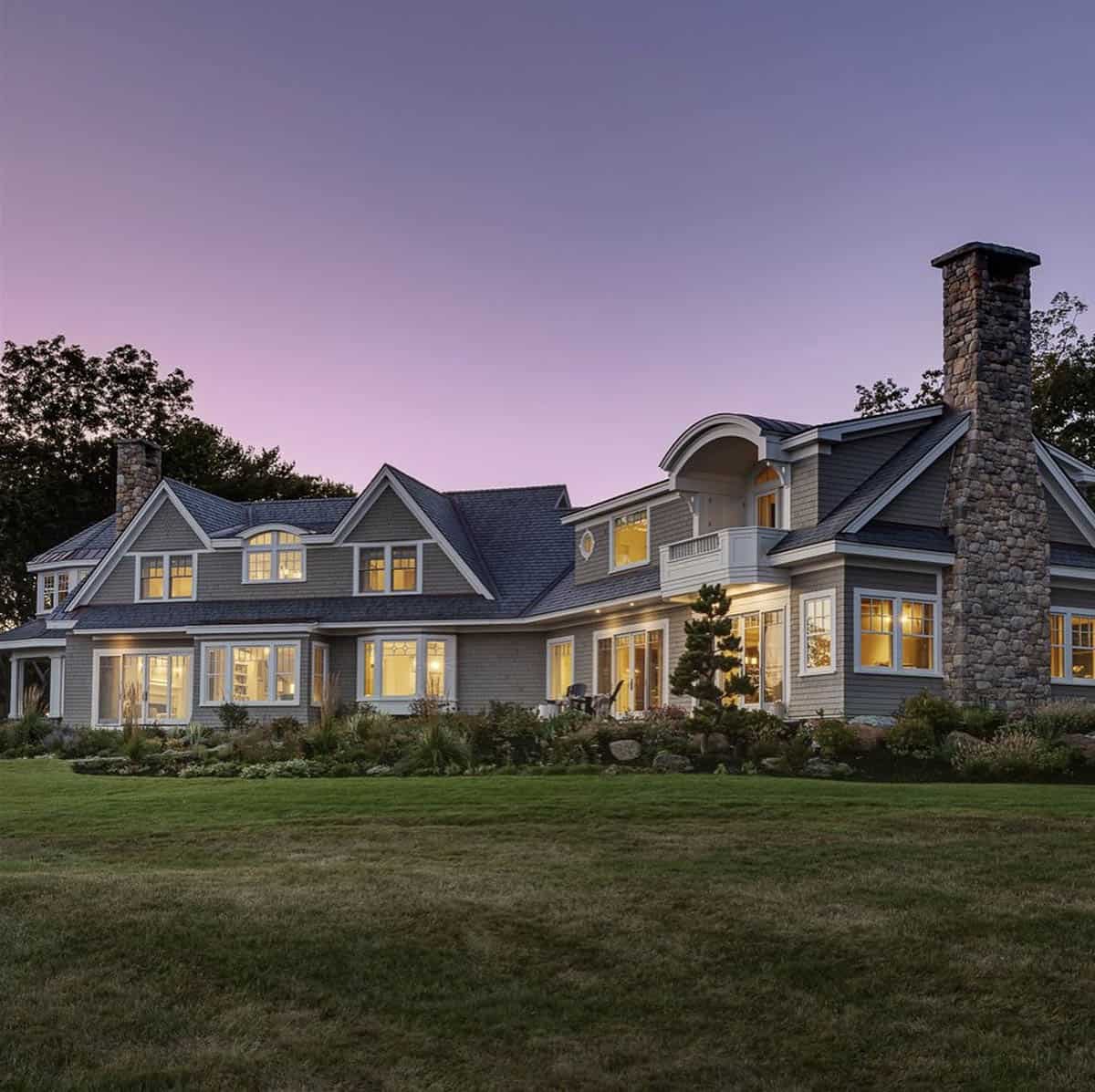Back of the house glowing at dusk under a purple sky, with a stone chimney, lit windows, and a landscaped slope