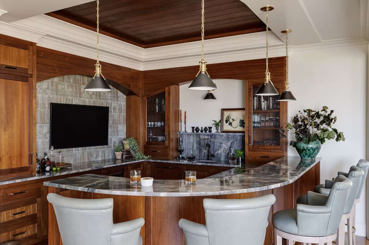 Home bar with a curved stone-topped counter, walnut cabinetry, brass pendants, and pale leather barstools