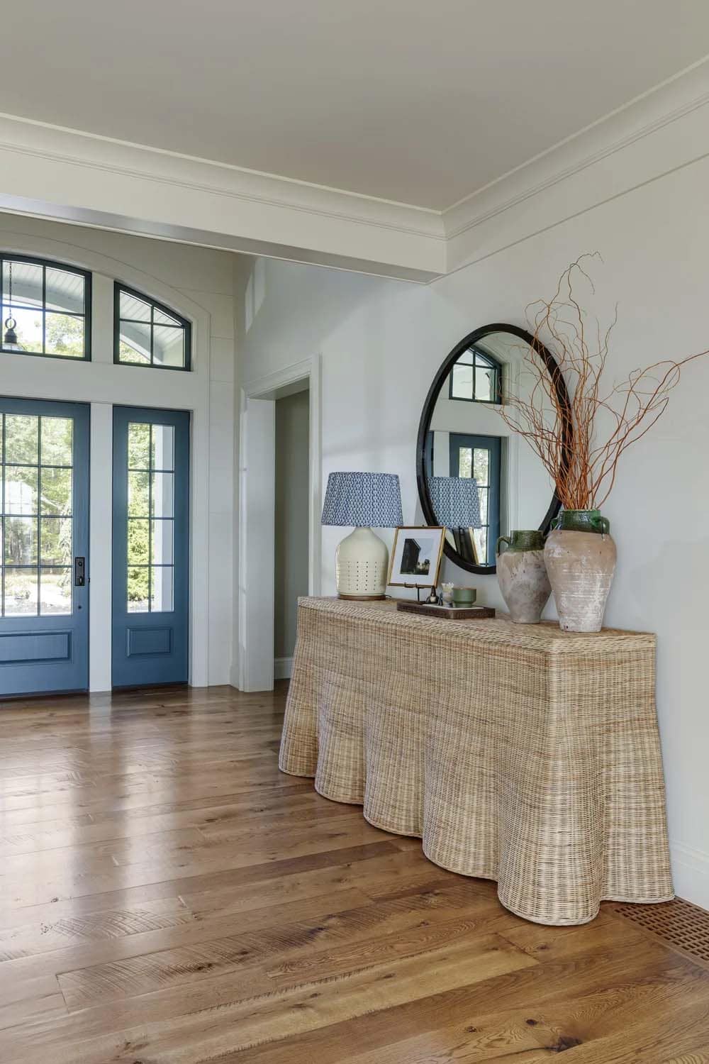 Foyer with a scalloped wicker console, a round black mirror, and blue double doors under an arched transom