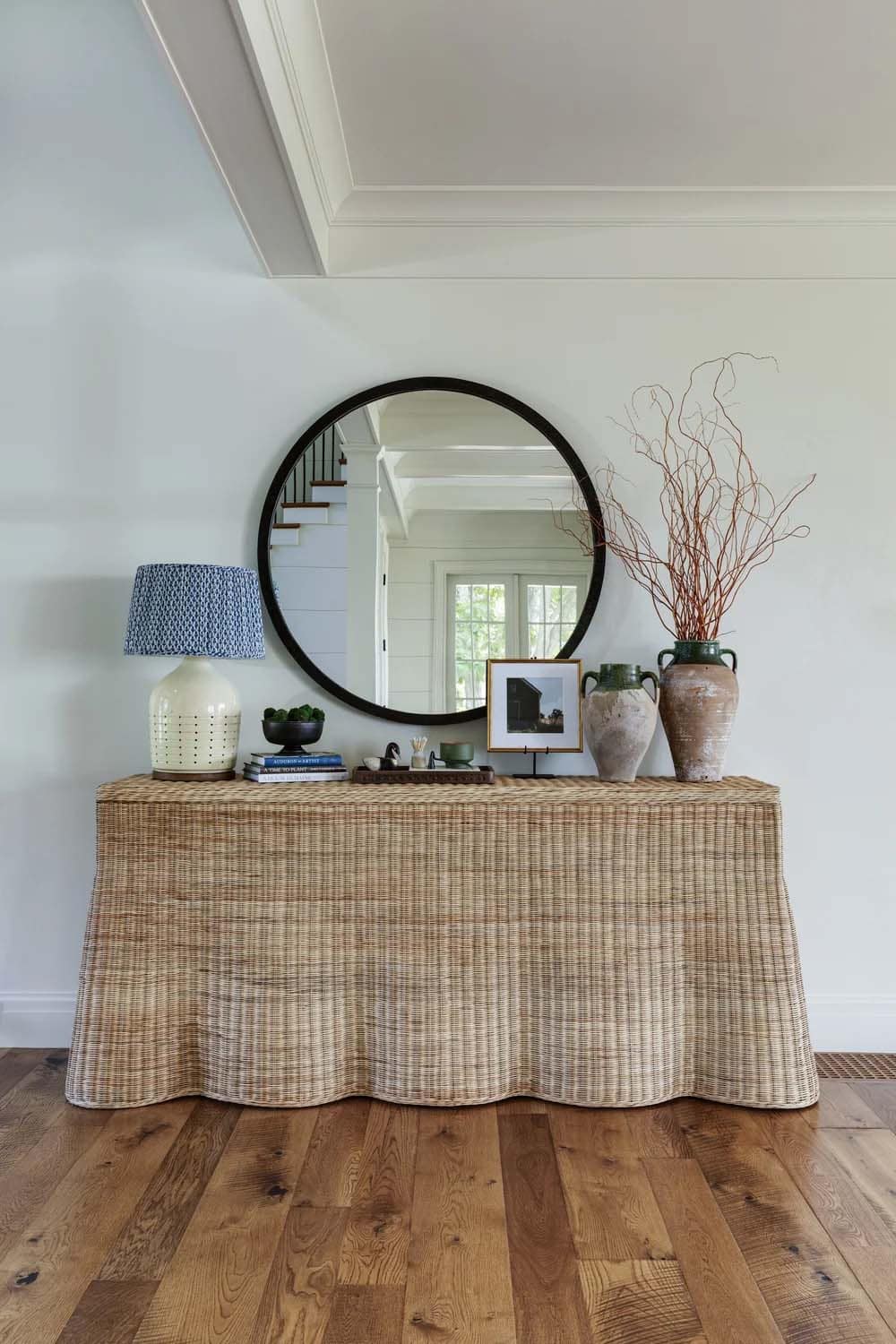 Close view of the wicker console with a round mirror, a patterned lamp, and ceramic vases holding bare branches
