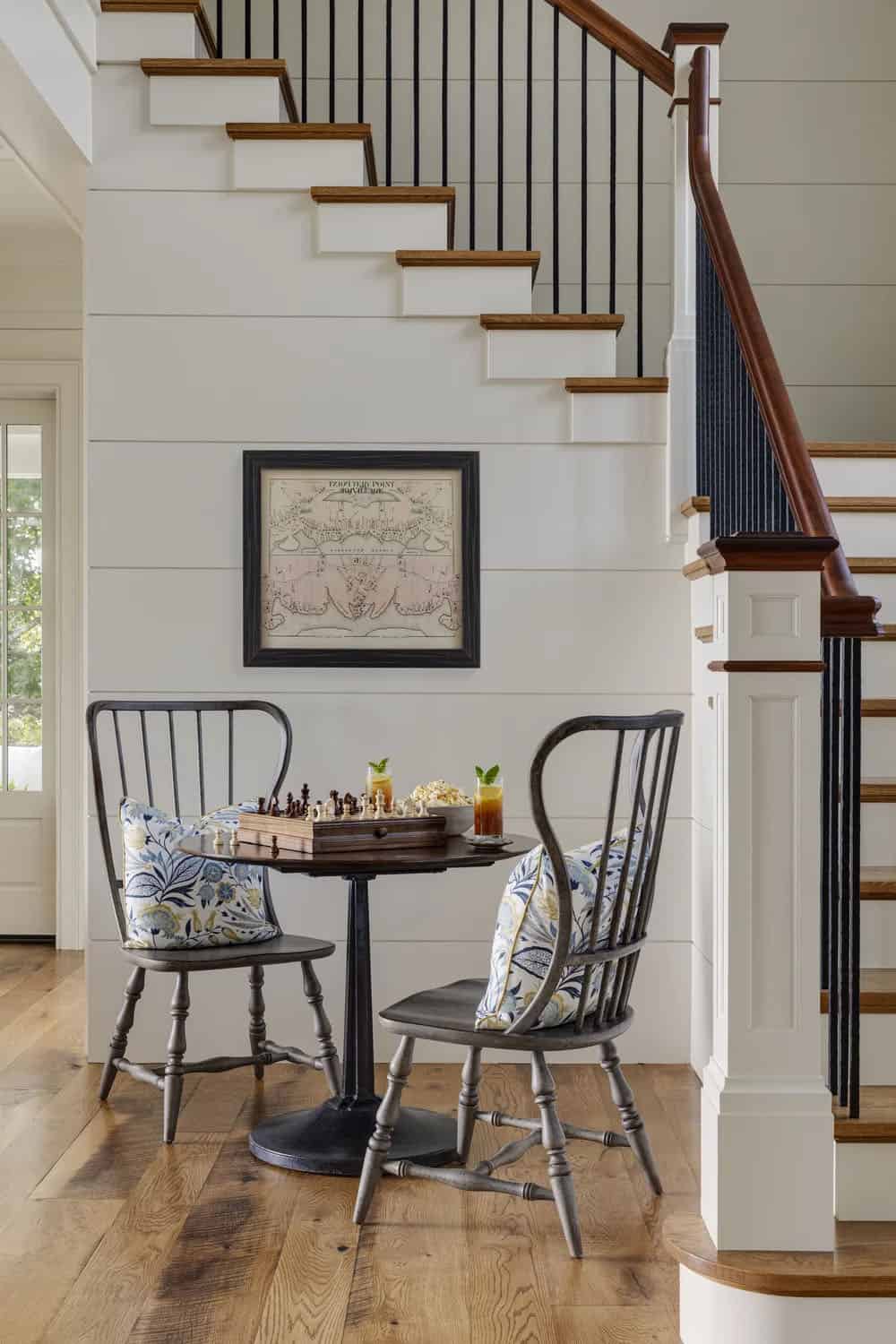 Chess table with two black Windsor-style chairs and patterned pillows beneath a shiplap staircase with a framed vintage map