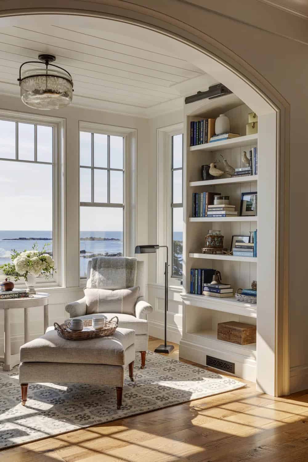 Reading nook seen through an arched opening, with an upholstered armchair and ottoman, built-in bookshelves, and ocean views through tall windows