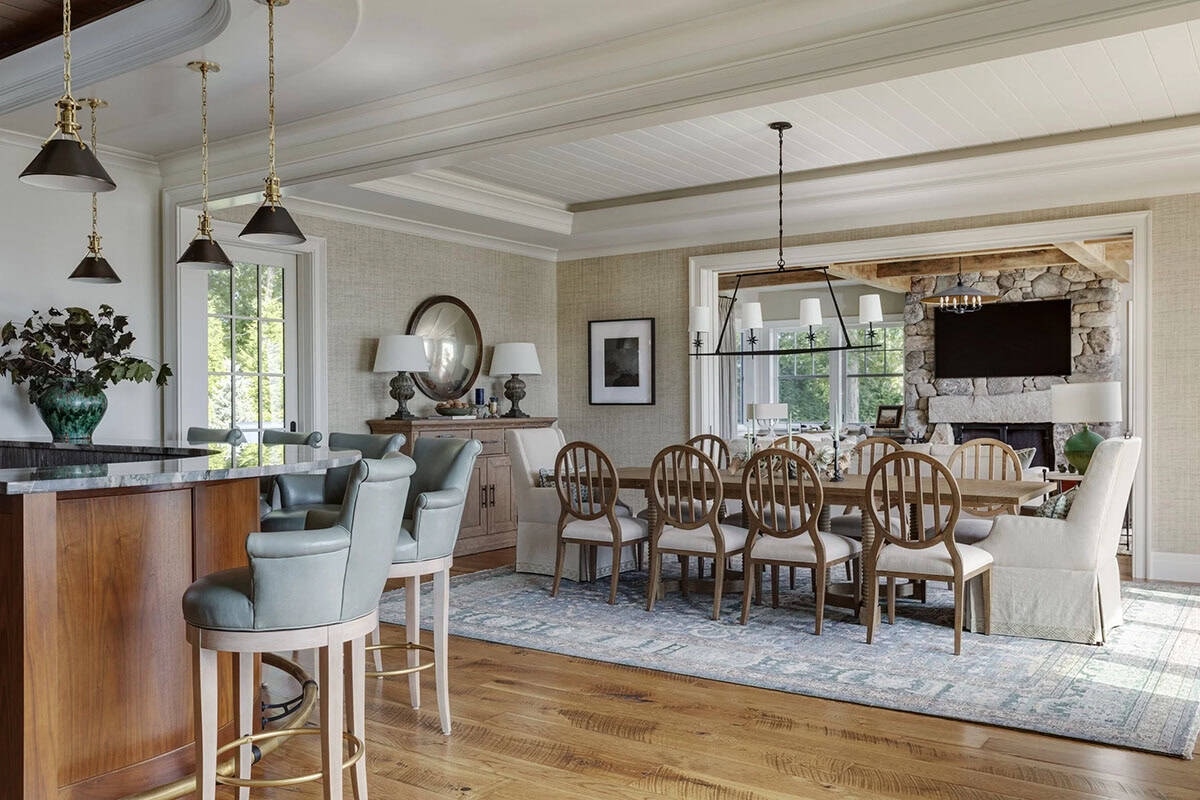Dining area with a long wood table and slat-back chairs, with the curved bar and leather barstools in the foreground and a stone fireplace beyond