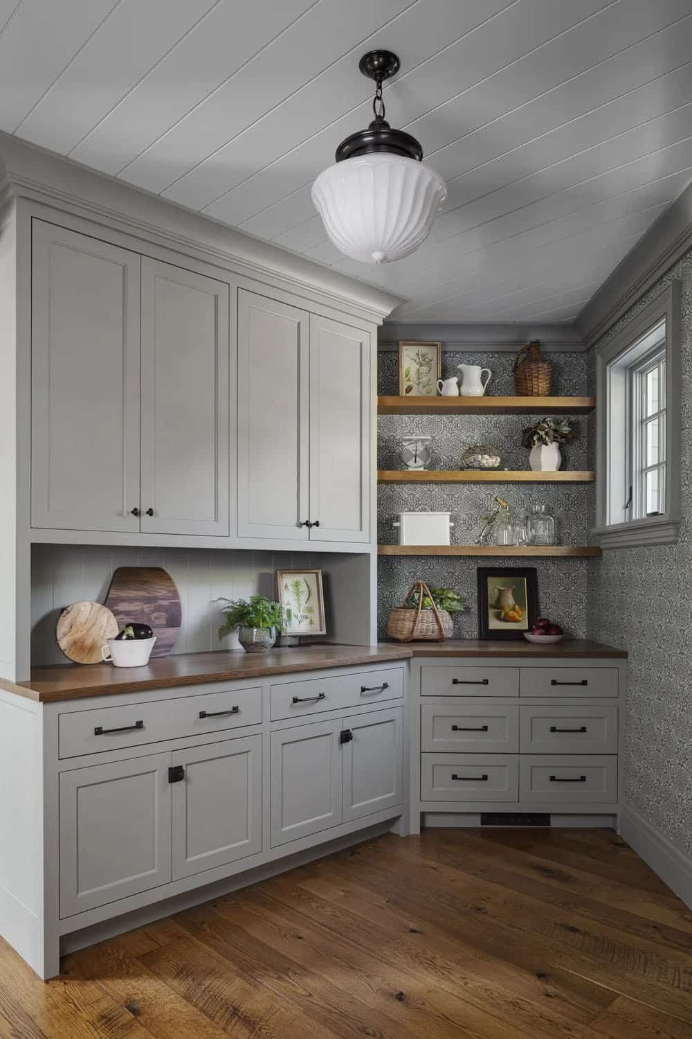 Pantry with gray-green cabinetry, a wood countertop, open oak shelves against patterned wallpaper, and a white schoolhouse pendant