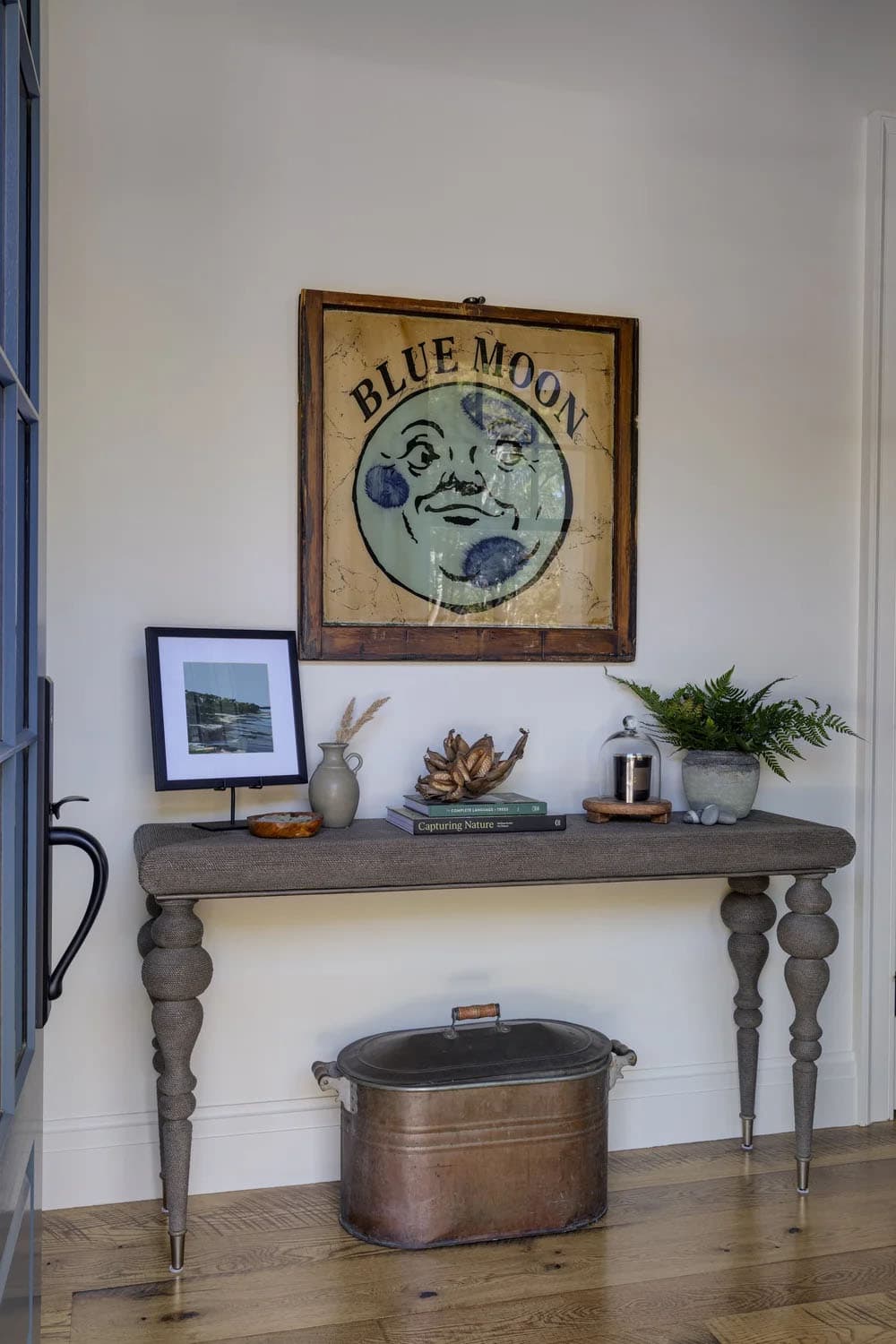 Rope-wrapped console table with turned legs, a vintage "Blue Moon" sign, a copper tub below, and a small display of plants and framed art