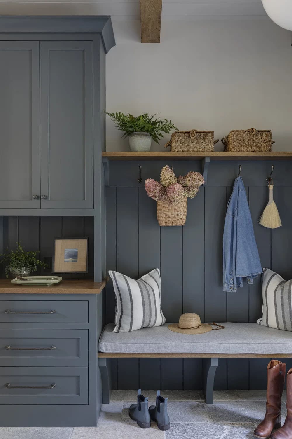 Mudroom bench with slate-blue paneling, wall hooks, a wood shelf with wicker baskets, and boots on a stone floor
