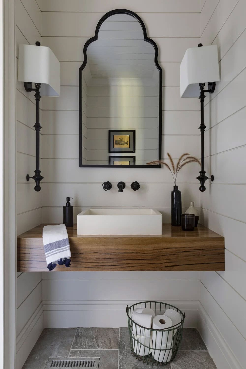 Powder room with shiplap walls, a floating wood vanity, a white vessel sink, an arched black mirror, and tall black sconces