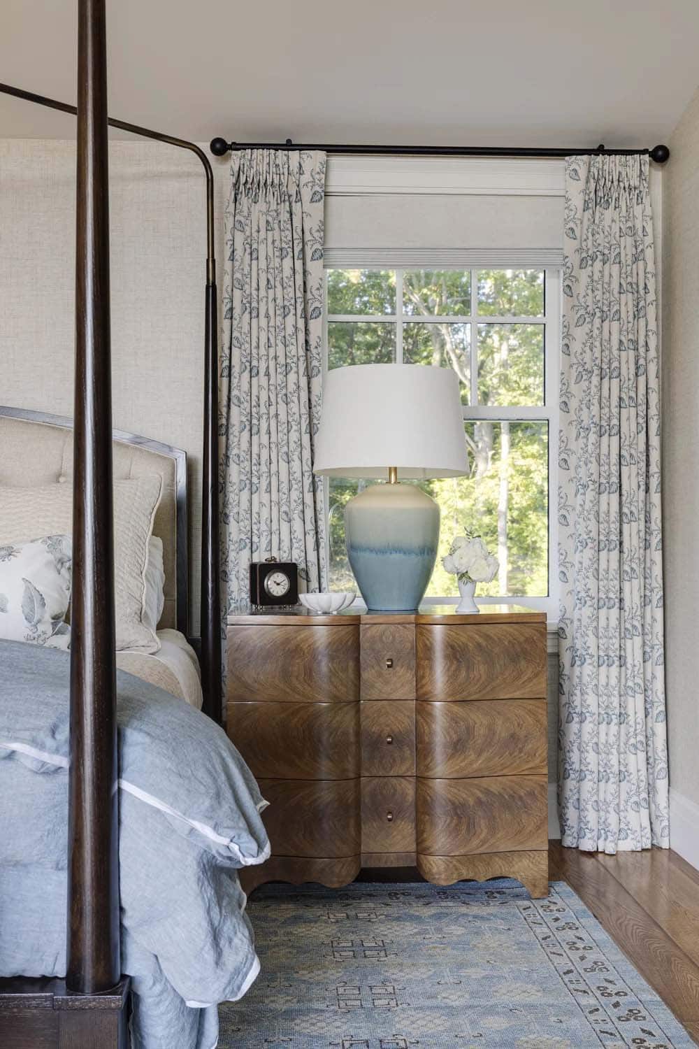 Bedside chest with curved burl-wood drawers, a blue ombré ceramic lamp, and floral curtains beside a four-poster bed