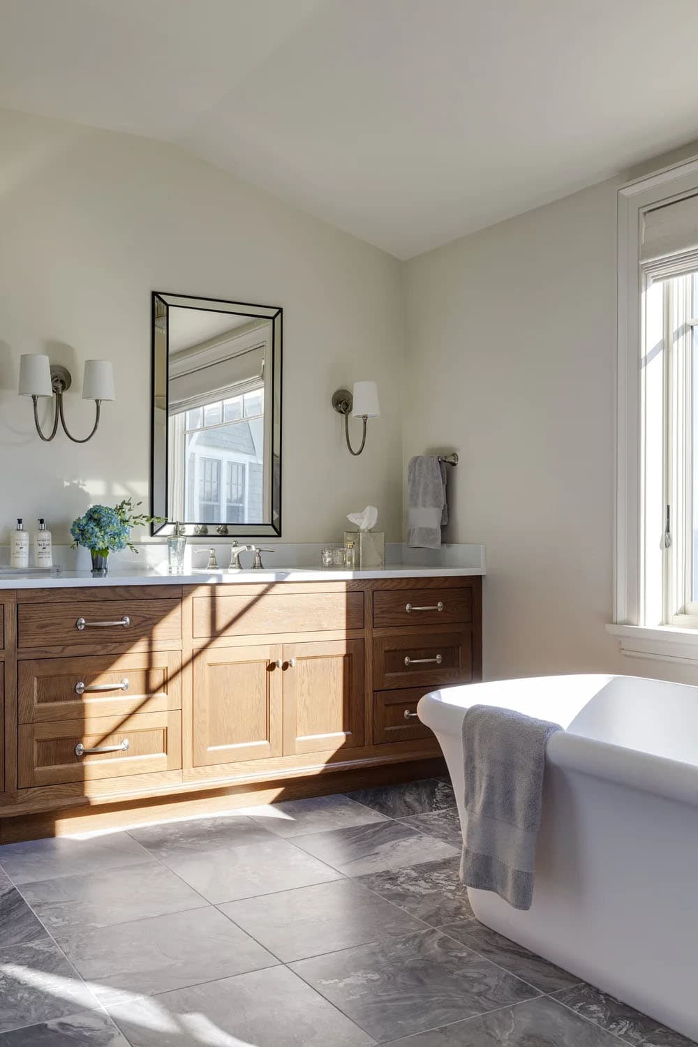 Primary bathroom with a long oak vanity, a beveled mirror, nickel sconces, a freestanding tub, and gray stone floor tile