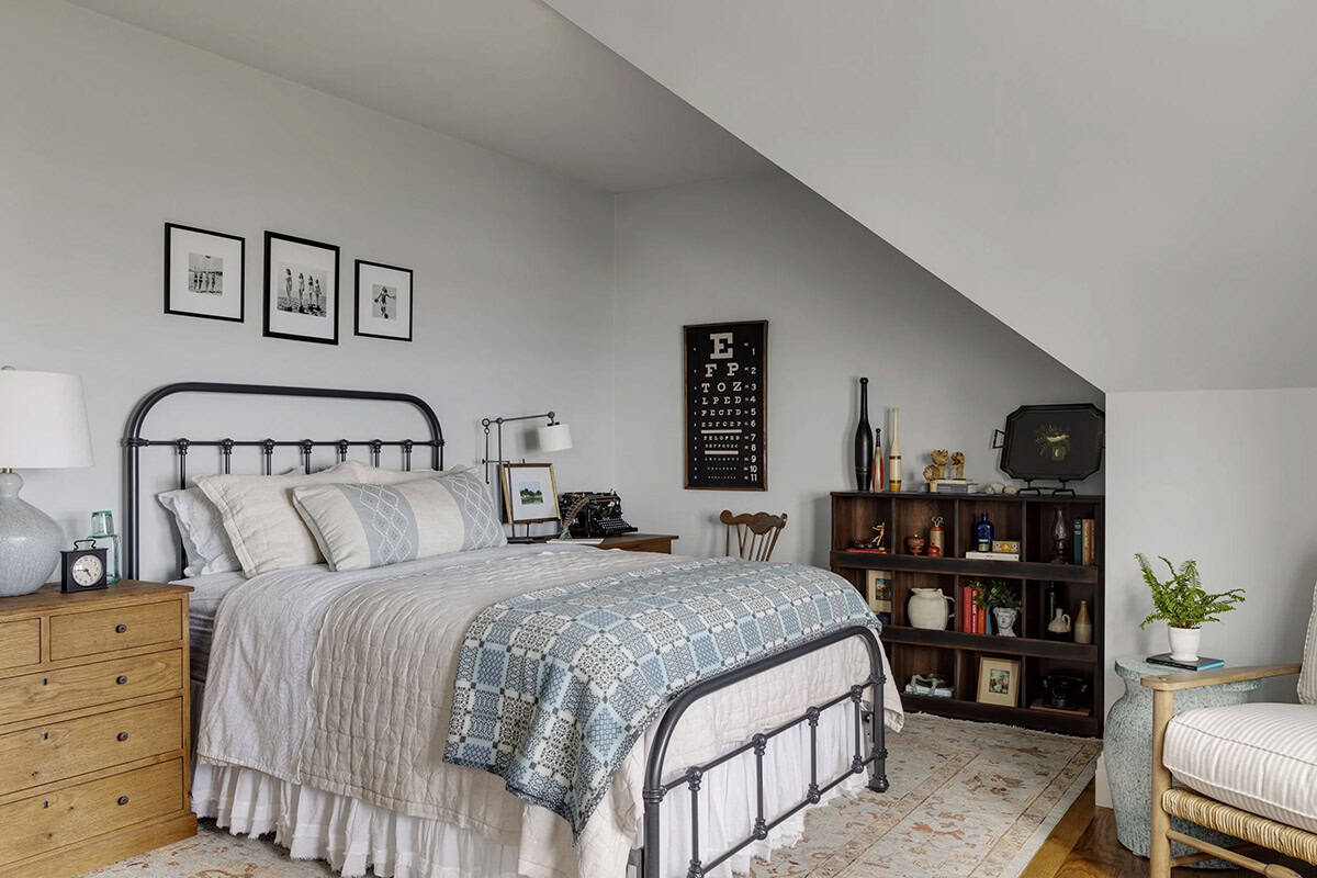 Guest bedroom under a sloped ceiling with an iron bed, a vintage eye chart, an open wood shelf unit, and a woven blue-and-white coverlet