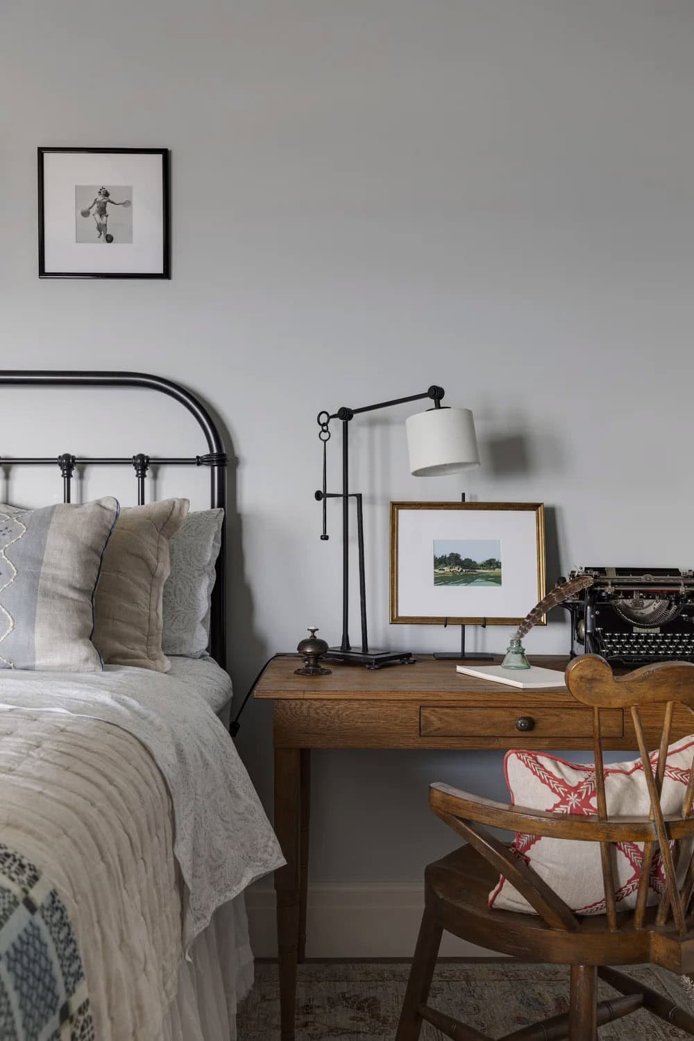 Bedside writing desk with a vintage typewriter, an adjustable black lamp, a framed landscape, and a wooden armchair