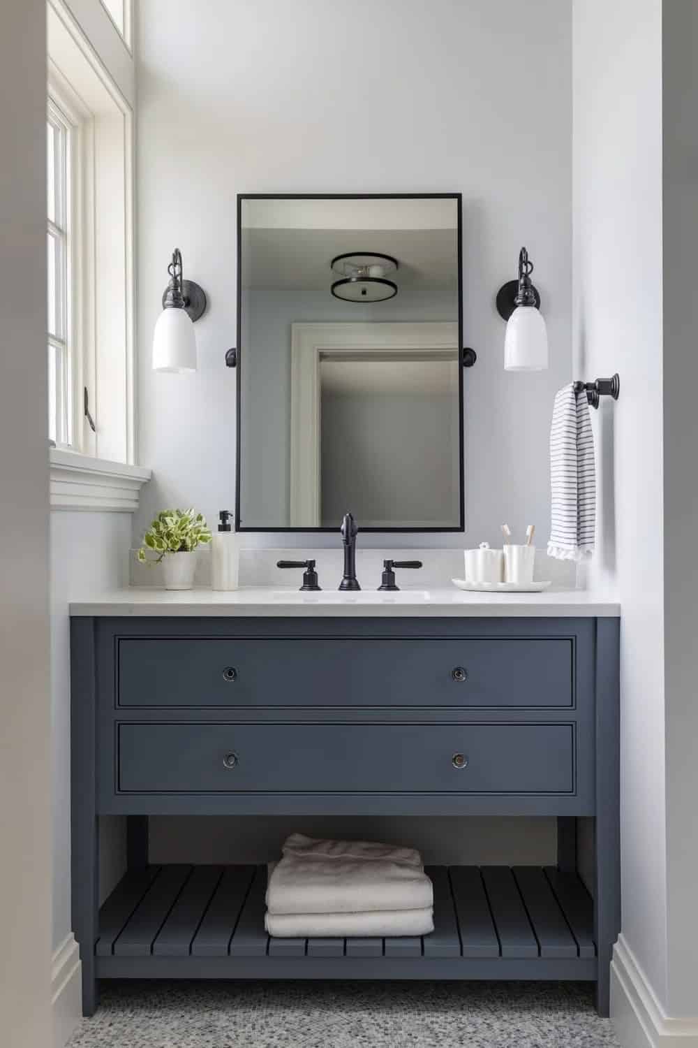 Bathroom vanity with slate-blue cabinetry, an open slatted shelf, a black-framed mirror, and white glass sconces