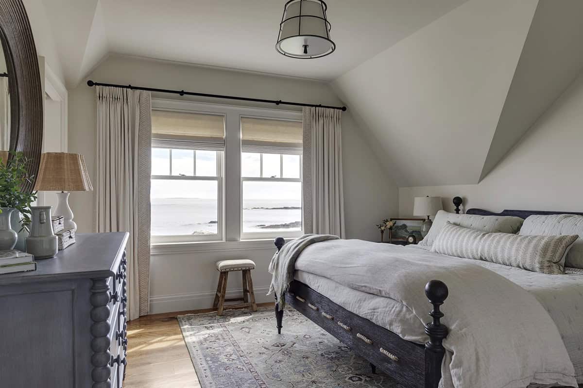 Bedroom with a dark four-poster bed, a gray-blue dresser, and two windows overlooking the ocean beneath a sloped ceiling