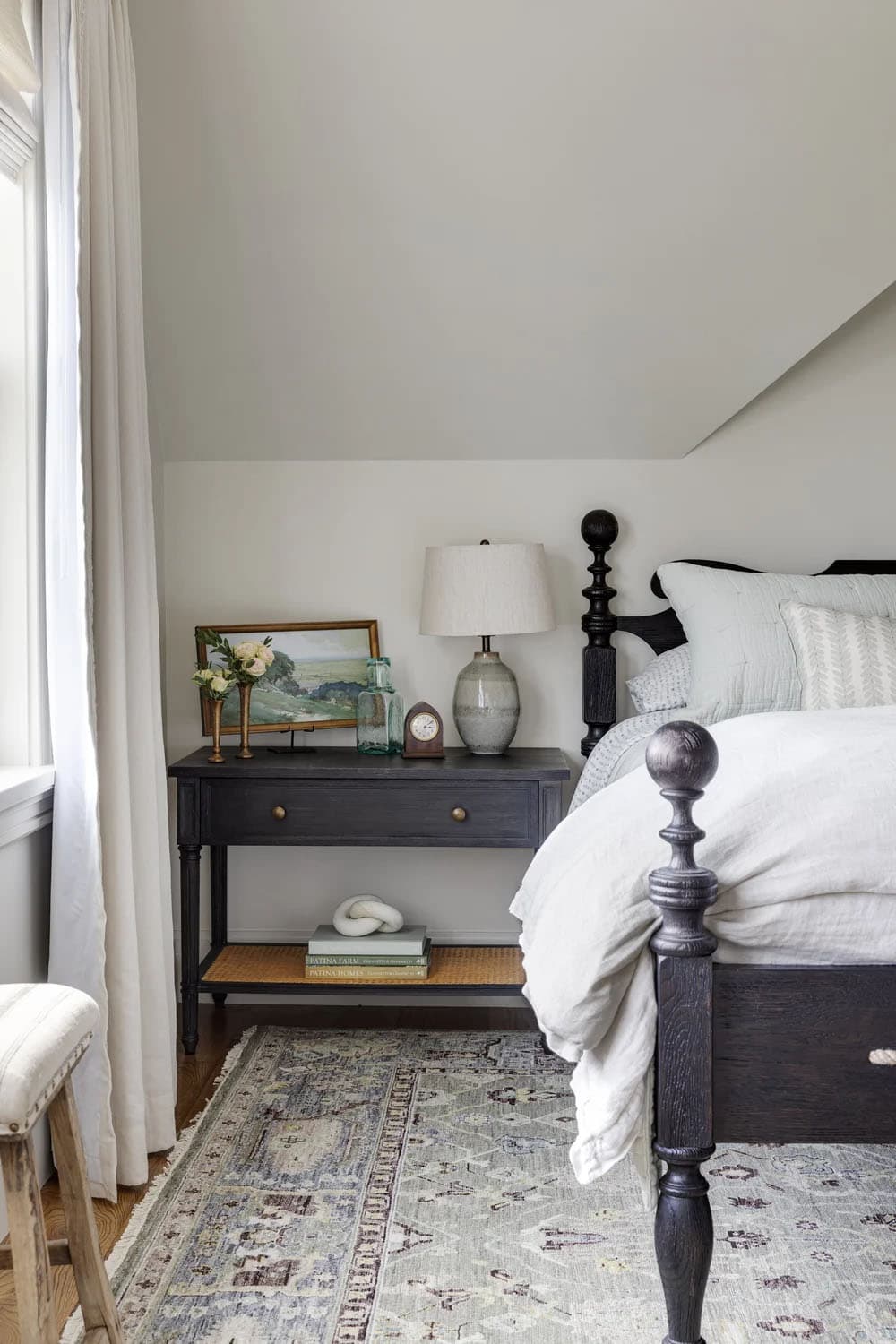 Bedside table with a ceramic lamp, a framed landscape, brass bud vases, and a dark carved bedpost on a patterned rug