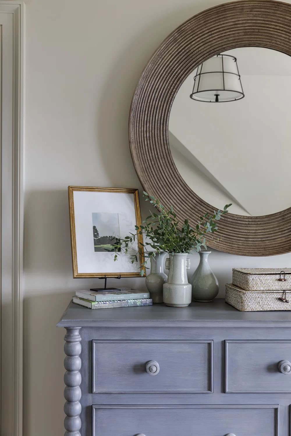 Gray-blue spindle-leg dresser with a large round ribbed mirror, celadon vases, eucalyptus stems, and woven lidded boxes