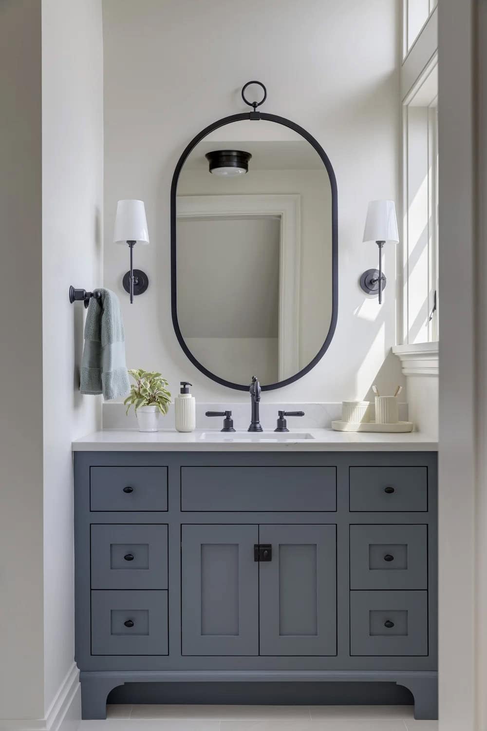 Bathroom vanity with blue-gray cabinetry, an oval black mirror, black sconces, and a window letting in sunlight