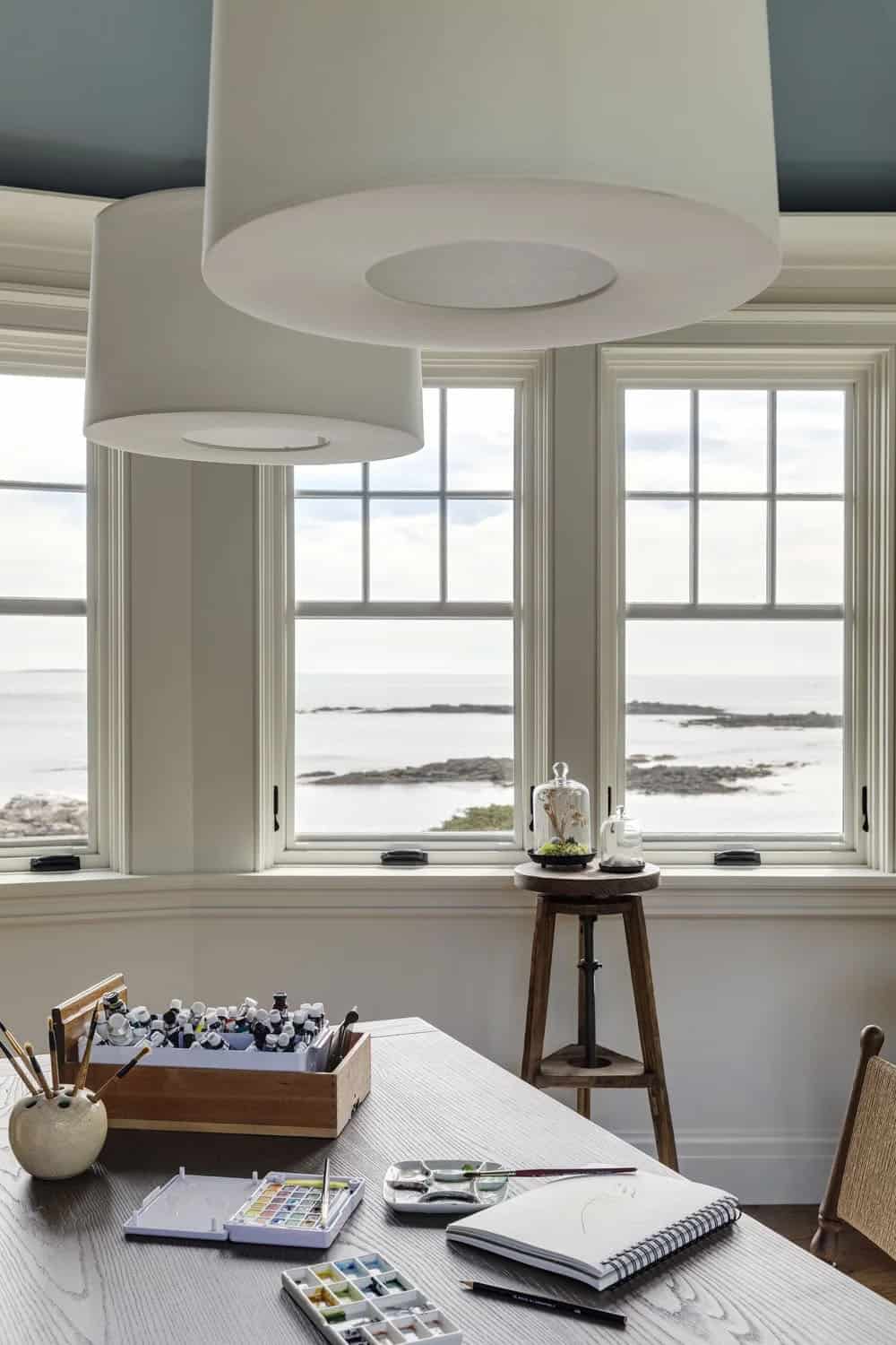 Art studio table with paint tubes, watercolor palettes, and a sketchbook, with a wooden stool and ocean views through the windows