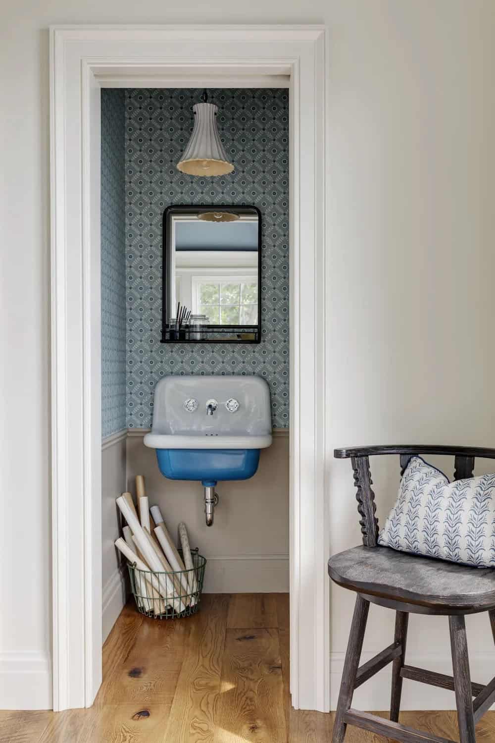 Small powder nook with blue patterned wallpaper, a wall-mounted blue-and-white sink, a black-framed mirror, and a basket of rolled papers