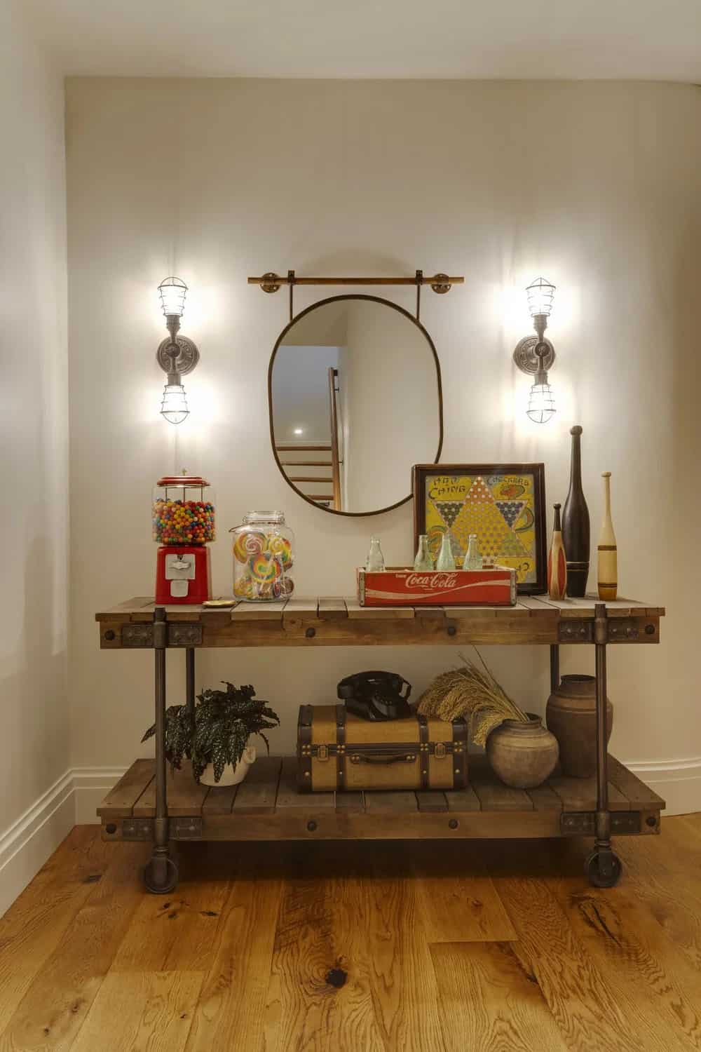 Rolling industrial console with a gumball machine, candy jars, a vintage Coca-Cola crate, and a Chinese checkers board beneath an oval mirror and caged sconces
