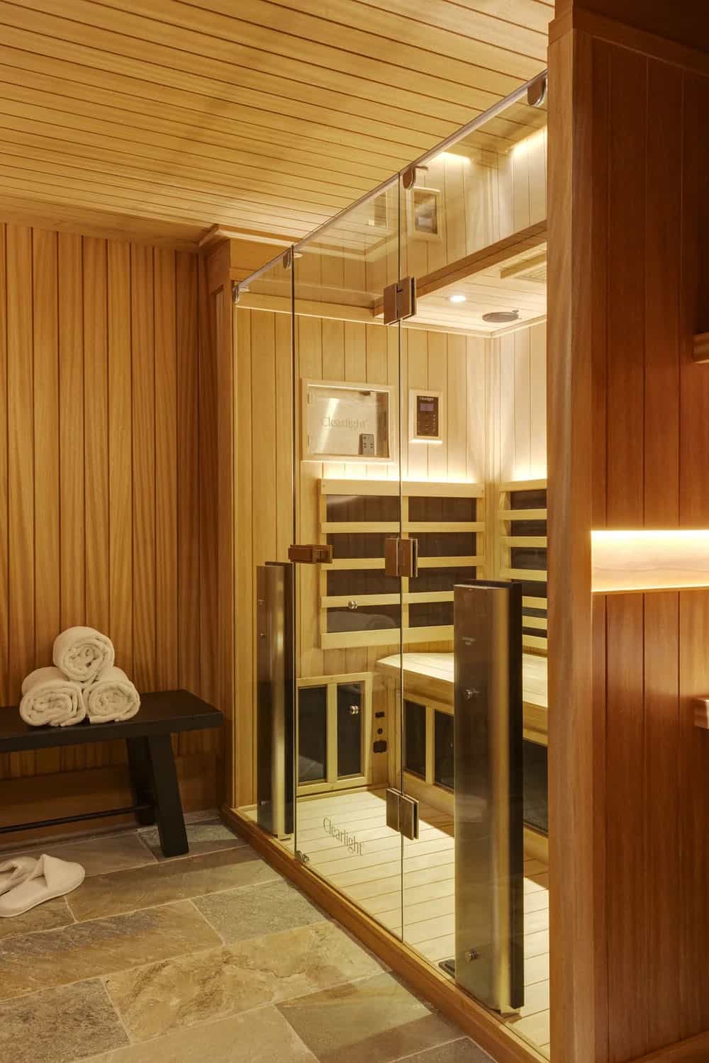 Infrared sauna with a glass front, light wood paneling, a dark bench holding rolled towels, and stone floor tile
