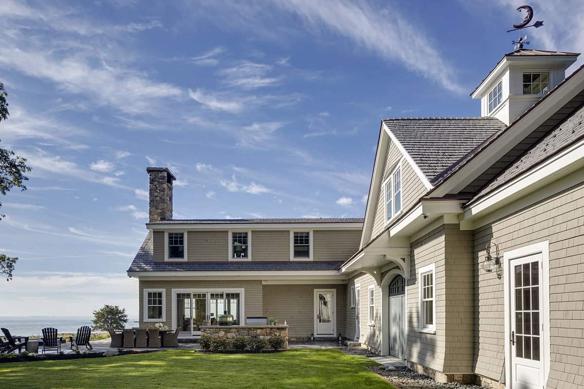 Rear of the house with a stone chimney, shingled wings, a patio with outdoor dining and Adirondack chairs, and a cupola with a moon weathervane