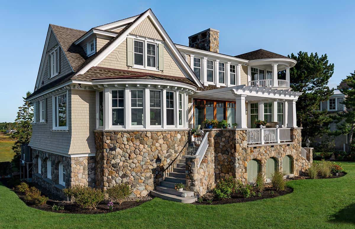Shingle-style coastal home with stone base, curved bay windows, and white pergola on a green lawn